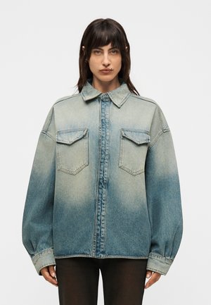 Γυναίκα που φοράει πολύ μεγάλη ξεθωριασμένη μπλε τζιν μπουφάν με μπροστινές τσέπες με καπάκια, στέκεται μπροστά σε απλό λευκό φόντο, κοιτάζοντας μπροστά.