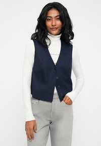 Kobieta z czarnymi falowanymi włosami, ubrana w białą golfową bluzkę, granatową kamizelkę zapinaną na guziki i jasnoszare dżinsy, pozująca z jedną ręką w kieszeni.