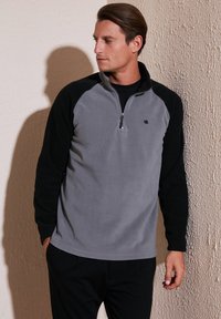 Fleece-pullover med grå krop og sorte ærmer, halv-lynlås foran og lille logo på brystet. Blød tekstur og afslappet pasform.
