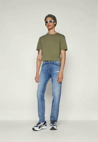 Ελιόχρωμο κοντομάνικο μπλουζάκι, ανοιχτό μπλε τζιν με φθορές, λευκά και μαύρα αθλητικά παπούτσια, γκρι σκούφος και λευκά γυαλιά ηλίου.