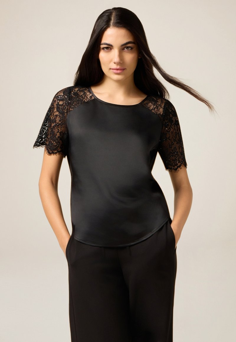 Czarna bluzka z satynowym korpusem i koronkowymi krótkimi rękawami ze ząbkowanym brzegiem. Gładka faktura, luźny krój i okrągły dekolt.