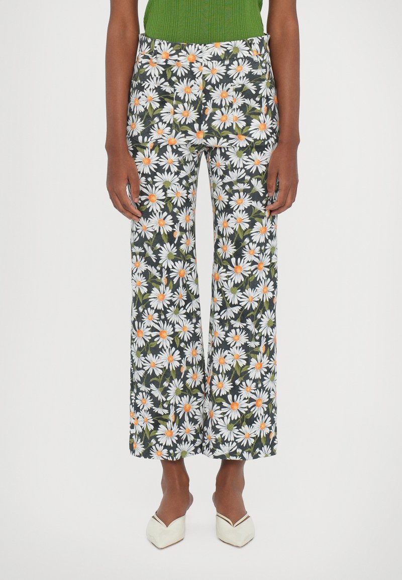 Femme portant un pantalon noir à imprimé marguerites blanches et orange, un haut vert et des chaussures blanches à bout pointu, debout sur un fond blanc.