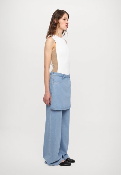Weißes geripptes ärmelloses Top mit beigen seitlichen Einsätzen, kombiniert mit weit geschnittenen, hellblauen Jeans mit einem vorderen Denim-Rocküberzug. Schwarze Schuhe.