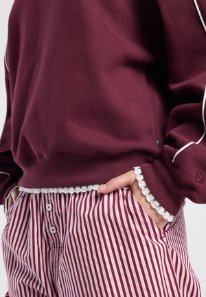 Bordeauxrode trui met witte bies en kanten afwerking bij de zoom en manchetten, gecombineerd met rood-wit gestreepte broek met knoopsluiting.