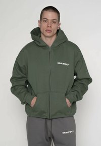 Grøn hættetrøje med lynlås og frontlomme, med hvidt "MULTIPLY"-logo. Blødt stof, løs pasform og stor hætte. Båret over grå joggingbukser.