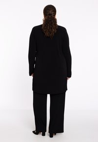 Schwarzes, geripptes Langshirt mit langen Ärmeln und passenden, weit geschnittenen Hosen, die einen geraden Schnitt und eine glatte Textur aufweisen. Pumps sind sichtbar.