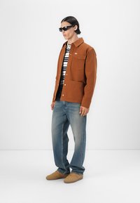 Bruine stoffen jas met een kraagontwerp, gecombineerd met een gestreept shirt, blauwe spijkerbroek en beige sneakers. Zonnebril maakt de look compleet.