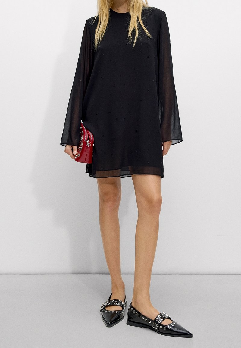 Robe mini noire transparente avec de longues manches évasées, associée à des ballerines noires à bout pointu avec des accents d'œillets argentés et un sac à main rouge.