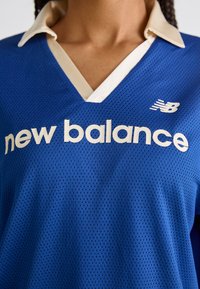 Kék hálós sportpóló, krémszínű gallérral és nagy fehér felirattal "new balance", bal oldalon logóval.
