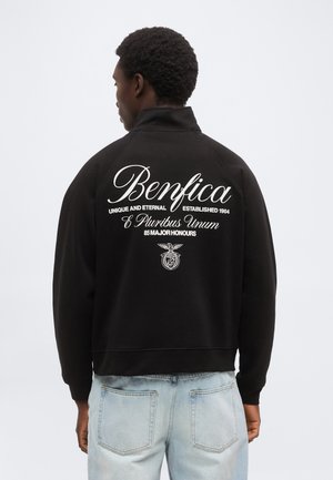 Άνδρας φοράει μαύρο μπουφάν με το κείμενο «Benfica», το μότο και το έμβλημα στην πλάτη, σε συνδυασμό με ανοιχτά μπλε τζιν, στραμμένος με την πλάτη προς την κάμερα.