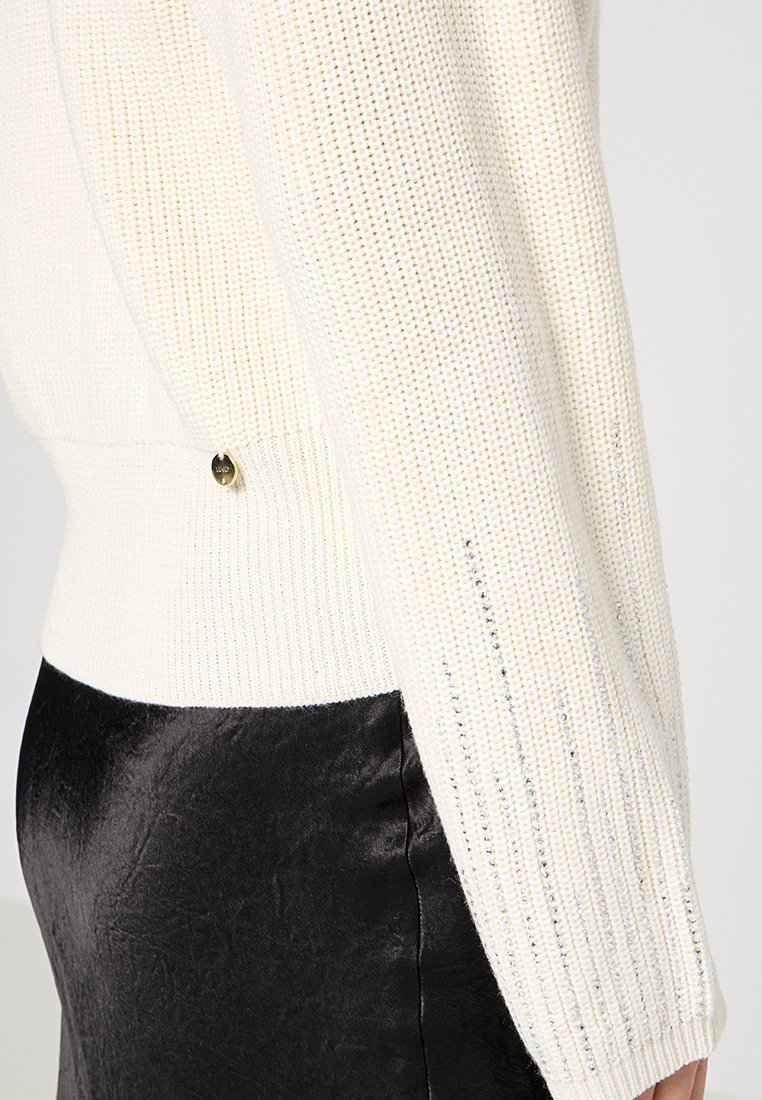 Maglione bianco a maglia con una texture a coste, caratterizzato da una vita aderente e lievi dettagli lucidi. Include un bottone color oro sul lato.