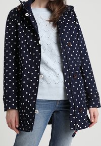 Marinblå regnjacka med vita prickar, utrustad med huva, knappstängning och justerbar dragsko i nederkant. Bärs över en ljus tröja.