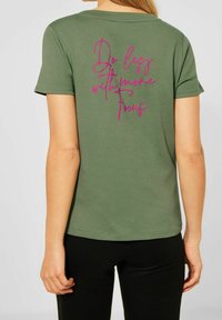 Olivgrön t-shirt med kort ärm och mjuk textur. Har en rosa inskrift på baksidan: "Gör mindre med mer fokus."