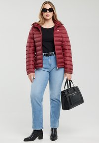 Žena v černých slunečních brýlích, červené péřové bundě, černém topu, světle modrých džínách a černých botách, držící černou kabelku, na šedém pozadí.