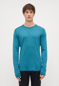 Camisa de manga longa e gola redonda em tecido verde-água, com costuras planas e punhos alongados, combinada com calças pretas. Textura suave.