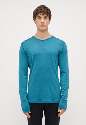 Camisa de manga longa e gola redonda em tecido verde-água, com costuras planas e punhos alongados, combinada com calças pretas. Textura suave.