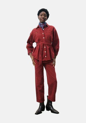 Rode corduroy jasje met riem, knoopsluiting en twee voorzakken, gedragen met bijpassende rode broek en zwarte enkellaarzen.