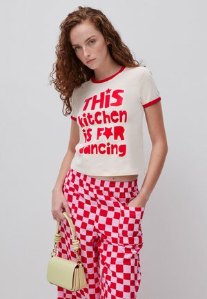 Vrouw met krullend haar, gekleed in een wit-rood graphic T-shirt met de tekst "Deze keuken is om te dansen" en rood-witte geruite broek, met een gele tas in haar hand.