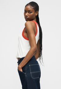 Žena s dlhými vrkočmi, oblečená v bielom bezrukávovom topu s oranžovým lemom a tmavomodrých nohaviciach s veľkými zadnými vreckami, pózujúca zboku.