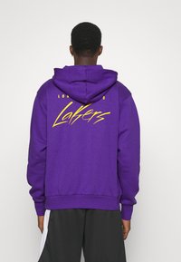 Sweat à capuche violet avec un grand logo jaune "Lakers" au dos, doté d'une capuche à cordon de serrage et de poignets et ourlet côtelés. Texture de tissu lisse.