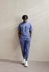 Niebieska bawełniana bluza z kapturem i dresy, z małym czarnym logo na klatce piersiowej i nogawkach. W połączeniu z białymi trampkami, stojąca na tle neutralnej ściany.