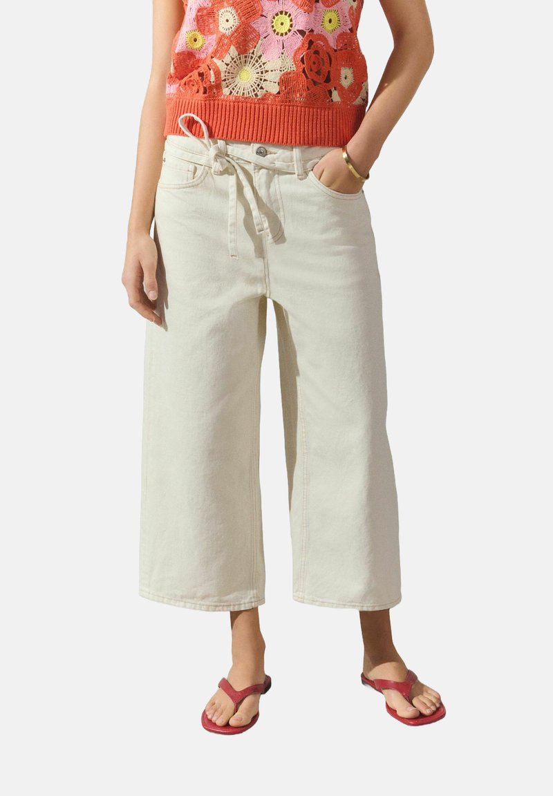 Pantalones de mezclilla cremosos de pierna ancha con una cinturilla ajustable, que presentan un discreto detalle de costura. Combinados con chanclas rojas.