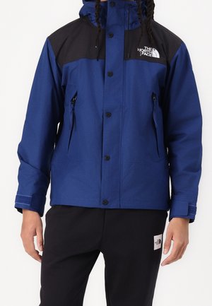 Man draagt een blauw-zwart gewatteerd jack met capuchon en ritssluitingen en een zwarte broek, beide met het logo van The North Face.