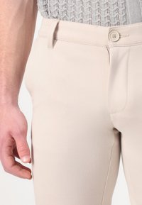 Pantaloni bej, croiți, cu o textură fină, închidere cu un singur buton și buzunare laterale înclinate. Materialul are o calitate moale și elastică.
