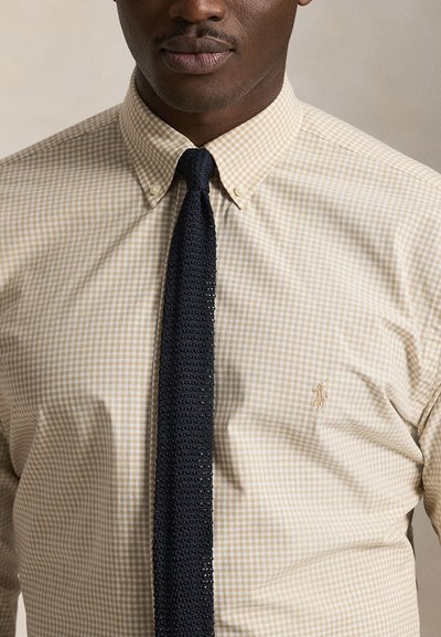 Homme portant une chemise beige à carreaux avec logo polo brodé et cravate en maille noire, debout devant un fond uni.