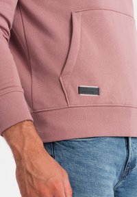Nahaufnahme einer Person, die ein rosafarbenes Sweatshirt mit Fronttasche und blaue Jeans trägt, mit einem schwarz-weißen Markenlabel auf der Tasche.