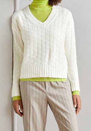 Personne portant un pull à col en V texturé blanc par-dessus un col roulé vert citron et un pantalon beige à fines rayures, sur fond gris clair.