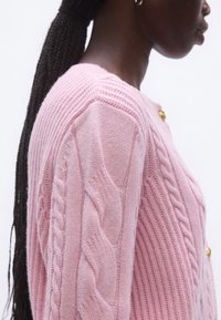Világos rózsaszín kötött pulóver bordázott és fonott mintákkal, kör alakú nyakkivágással és egy díszes arany gombbal a bal vállon.