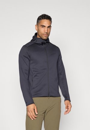 Donkergrijze zip-up hoodie met hoge kraag, gestructureerde stof en zijzakken, gecombineerd met lichtbruine broek.