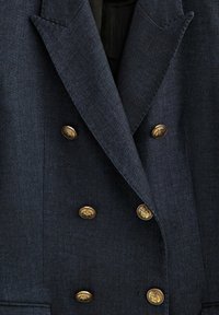 Blazer croisé bleu marine foncé avec six boutons dorés embossés et larges revers à pointe, montrant le détail du torse avant.