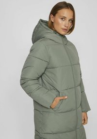 Manteau rembourré vert clair avec capuche, dotée d'une fermeture éclair sur le devant, de poches latérales et de panneaux horizontaux cousus pour la texture.
