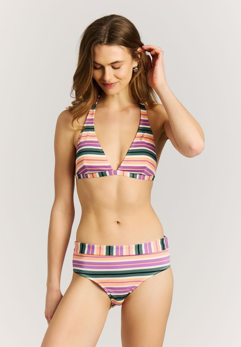 Kobieta poprawiająca włosy, ubrana w wielokolorowy, paskowany bikini z topem na szyję i majtkami z wysokim stanem na tle jednolitego tła.