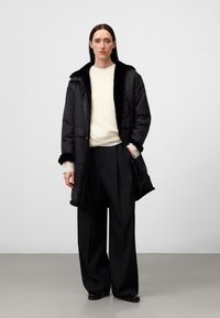 Kvinna med långt mörkt hår bär en svart furfoderjacka, vit tröja, svarta vida byxor och svarta skor, stående mot en enkel bakgrund.