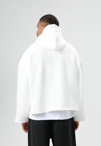 Sweat à capuche blanc surdimensionné avec cordon de serrage et grande capuche. Présente des épaules tombantes et un tissu doux. Porté sur une chemise blanche et un pantalon noir.