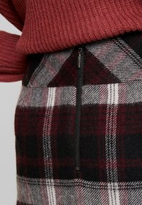 Ruudullinen hame, jossa on punaisia, mustia ja harmaita sävyjä sekä vetoketjusuljin. Kankaan pinta on pehmeä ja siinä on klassinen ruutukuosi.