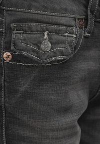 Jean en denim noir avec une petite poche avant, des détails usés, une quincaillerie en cuivre et des coutures contrastantes près de la ceinture.