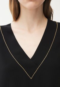 Mujer joven con blusa negra de cuello en V con un pequeño borde de cuentas doradas a lo largo del escote, enfocándose en el cuello y la parte superior del pecho.