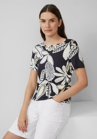 T-shirt à manches courtes bleu marine avec un motif de feuilles blanches, encolure ronde, tissu doux, associé à un short blanc. Le modèle se tient devant un fond clair.