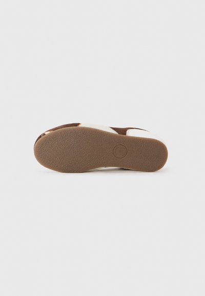 Calzature in suede marrone con punta arrotondata e accenti bianchi. Suola in gomma texturizzata con un dettaglio del logo circolare. Profilo piatto e design informale.