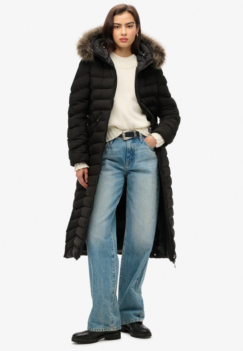 Abrigo tipo plumas negro con capucha forrada de pelaje, de largo completo, combinado con un suéter crema y jeans de pierna ancha en azul claro, terminado con botas negras.