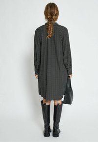 Schwarz gemustertes Kleid mit langen Ärmeln und hohem Kragen, hat einen lockeren Schnitt. Kombiniert mit knielangen schwarzen Stiefeln und einer kleinen schwarzen Tasche.