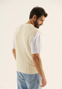 Pulover din tricot fără mâneci, de culoare crem, cu un model de cablu, purtat peste un tricou alb, asociat cu blugi de culoare albastru deschis.