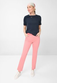 Marineblaues Kurzarmoberteil kombiniert mit rosa geraden Hosen und weißen Sneakers. Das Model trägt ein gemustertes Tuch und eine schlichte Halskette als Accessoires.