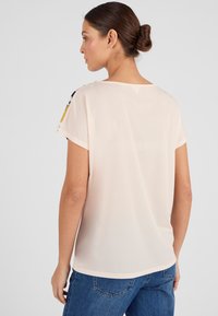 Mujer con el pelo castaño recogido en un moño, lleva una blusa de mangas cortas rosa claro y jeans azules, de espaldas contra un fondo claro y liso.