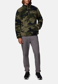 Pullover in pile con motivi camouflage, dotato di collo alto con zip e tasca sul petto, abbinato a pantaloni grigi e sneakers nere.