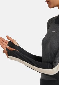 Vrouw in een grijs en zwart lange mouwtop met duimgaten, die haar handen naar voren uitstrekt tegen een effen achtergrond.